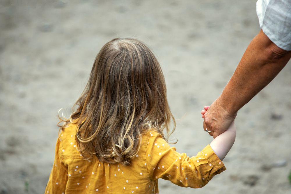 Liten jente med gul bluse som går med ryggen til kamera og holder en voksen person i hånda.